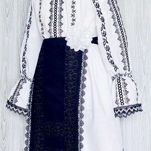 May include: White dress with black embroidery and a black skirt. The dress has long sleeves with flared cuffs, a round neckline, and a white flower at the waist. The skirt features a floral lace design.