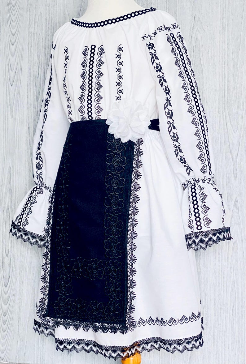 May include: White dress with black embroidery and a black skirt. The dress has long sleeves with flared cuffs, a round neckline, and a white flower at the waist. The skirt features a floral lace design.