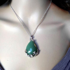 May include: A silver-toned necklace featuring a teardrop-shaped, green pendant. The pendant is accented with a decorative silver-colored floral design and small clear stones. The necklace is displayed on a mannequin.