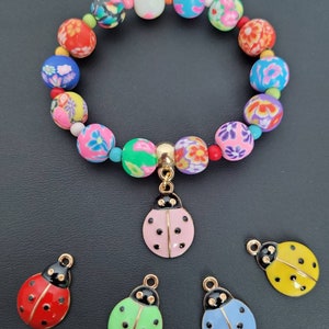 Op de afbeelding: Een kleurrijke kralen armband met een bloemenpatroon en een lieveheersbeestje bedel. De armband is gemaakt van polymer clay kralen en heeft een gouden sluiting. De lieveheersbeestje bedel is ook goudkleurig en heeft zwarte stippen.