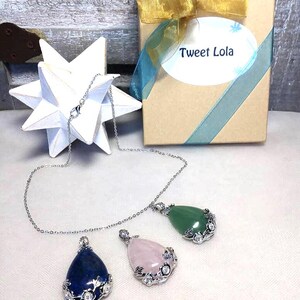 May include: Three gemstone pendant necklaces with silver chains. The pendants are teardrop-shaped, in blue, pink, and green. Each pendant is accented with a silver floral design. A gift box with a gold bow and the text "Tweet Lola" is in the background.