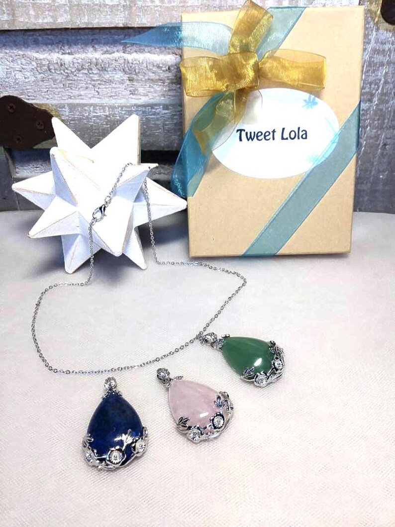 May include: Three gemstone pendant necklaces with silver chains. The pendants are teardrop-shaped, in blue, pink, and green. Each pendant is accented with a silver floral design. A gift box with a gold bow and the text "Tweet Lola" is in the background.