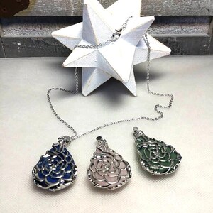 May include: Three gemstone pendant necklaces with silver chains. The pendants are teardrop-shaped with a floral design, in blue, pink, and green. A white star-shaped decoration is in the background.