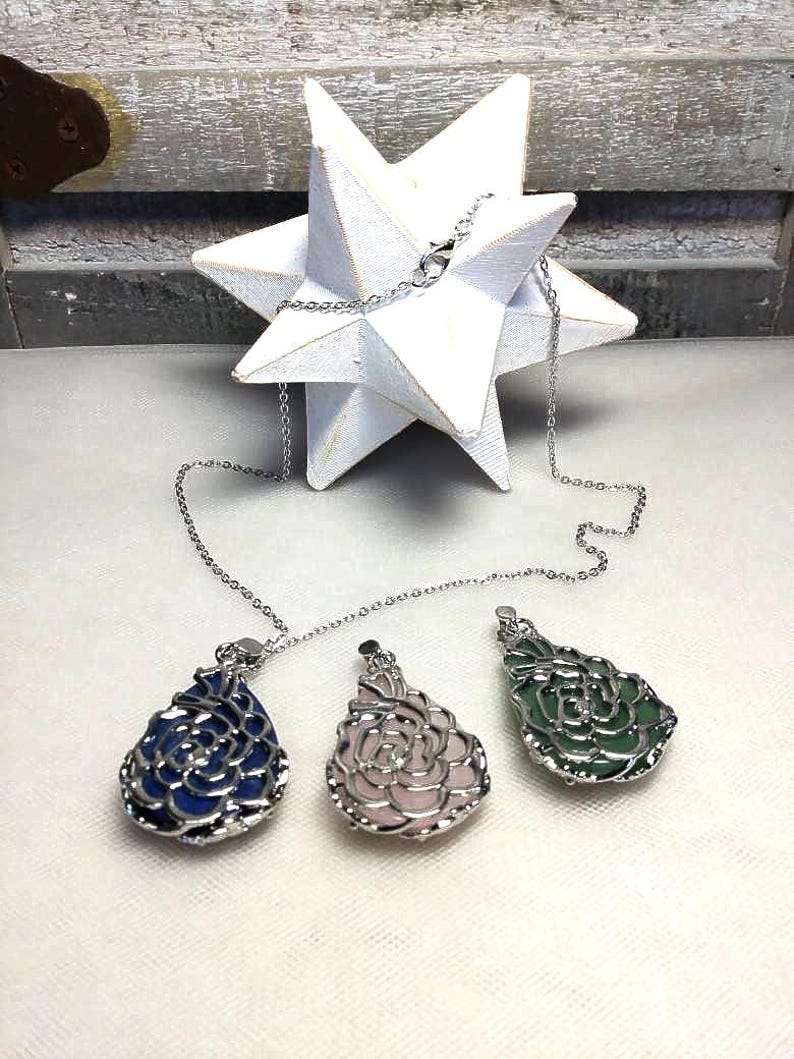 May include: Three gemstone pendant necklaces with silver chains. The pendants are teardrop-shaped with a floral design, in blue, pink, and green. A white star-shaped decoration is in the background.