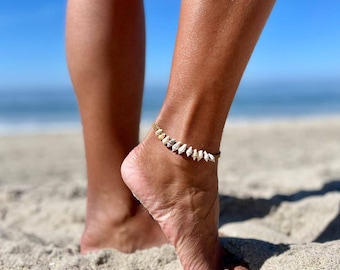 May include: A close-up of a person's ankle wearing a shell anklet. The anklet is made of small, white shells strung together on a gold chain. The person is standing on a sandy beach.