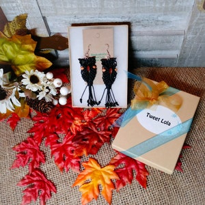 Op de afbeelding: Een paar zwarte en oranje macramé oorbellen met houten kralen, verpakt in een cadeaudoos met de tekst "Tweet Lola" op het label.