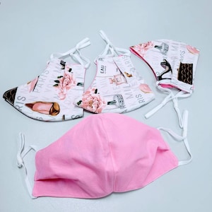 May include: Four face masks, three with a white background and a pink floral print with the text "Paris" and "New York" and one solid pink face mask.
