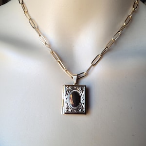 May include: Gold-toned necklace with a rectangular locket pendant. The locket features an oval, dark-colored center stone and ornate silver-colored detailing. The chain is a simple link style.