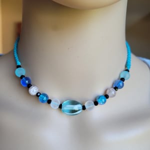 May include: A beaded necklace featuring a variety of blue, white, and black beads. The necklace has a turquoise cord and a large, oval-shaped, light blue bead in the center. Other beads include clear, frosted, and sparkly blue beads.