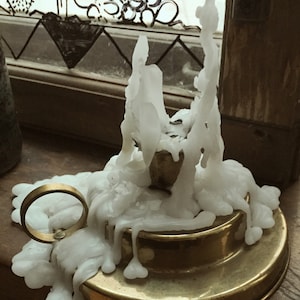 May include: A brass candle holder with a ring handle, covered in melted white wax. The wax has dripped down the sides of the holder, creating a textured appearance. The candle holder sits on a wooden surface.