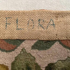 May include: Close-up of a beige fabric with a floral pattern. The fabric has a burlap border with the word "FLORA" written in blue ink.