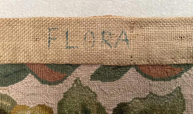 May include: Close-up of a beige fabric with a floral pattern. The fabric has a burlap border with the word "FLORA" written in blue ink.