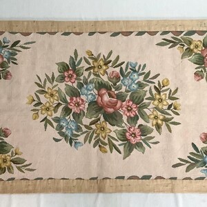 May include: A rectangular beige rug with a floral design. The rug has a brown border and features a central floral motif with pink, yellow, and blue flowers. The rug is made of a woven fabric and has a vintage aesthetic.