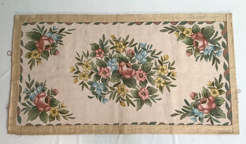 May include: A rectangular beige rug with a floral design. The rug has a brown border and features a central floral motif with pink, yellow, and blue flowers. The rug is made of a woven fabric and has a vintage aesthetic.
