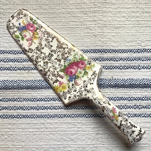 Puede incluir: Un servidor de pastel de cerámica blanca con un patrón floral y detalles dorados. El mango tiene forma de hoja y tiene un diseño floral.