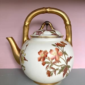 Peut inclure: Une théière en porcelaine blanche avec une poignée en bambou doré et un motif floral. La théière a un corps rond et un couvercle avec un motif floral. La théière est décorée de fleurs rouges, roses et vertes.