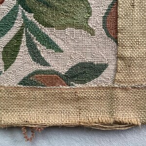 May include: Close-up of the bottom edge of a woven tapestry with a floral design. The tapestry has a brown burlap border and a beige background with green and brown floral patterns.