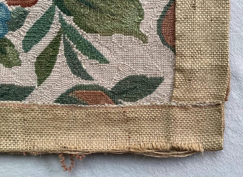 May include: Close-up of the bottom edge of a woven tapestry with a floral design. The tapestry has a brown burlap border and a beige background with green and brown floral patterns.