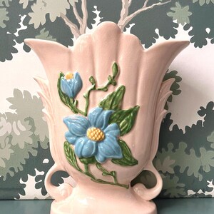 Może przedstawiać: Różowy ceramiczny wazon z falistą krawędzią i dwoma uchwytami. Wazon jest ozdobiony niebieskimi i zielonymi kwiatami w wypukłym wzorze.