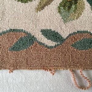May include: Close-up of the bottom edge of a hand-woven rug with a floral design. The rug is made of brown, green, and white yarn. The edge of the rug is frayed and has loose threads.