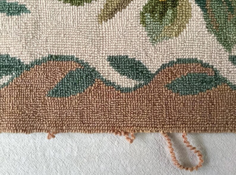 May include: Close-up of the bottom edge of a hand-woven rug with a floral design. The rug is made of brown, green, and white yarn. The edge of the rug is frayed and has loose threads.