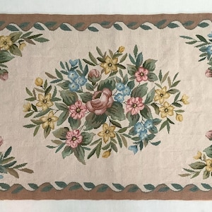 May include: A beige rug with a brown border and a floral design. The rug features pink, yellow, and blue flowers with green leaves.