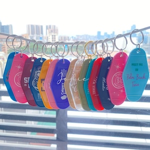 Puede incluir: Una colección de llaveros coloridos estilo motel colgando de un alambre. Los llaveros son de varios colores, incluyendo azul, rosa, rojo y amarillo, con texto y diseños. Uno de los llaveros dice "Meet Me in Palm Beach, Texas".