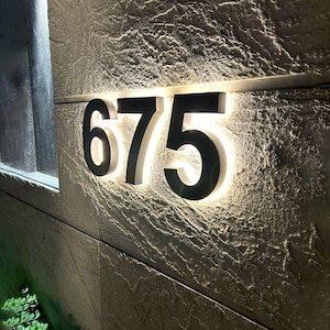 May include: Illuminated house numbers "675" in a modern font, mounted on a textured stone wall. The numbers are black with a bright white backlight, creating a striking contrast. The numbers are approximately 10 inches wide.