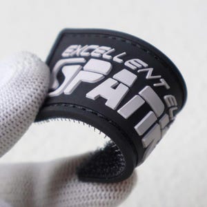 May include: A flexible black rubber patch with raised white lettering that reads EXCELLENT SPARK. The patch features a curved rectangular shape with a hook-and-loop fastener backing, designed for attachment to fabric surfaces or gear.