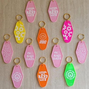 May include: A collection of colorful motel-style keychains. The keychains are in various colors, including pink, yellow, orange, and green. Some keychains have text that reads "Living My Best Life," "No Bad Days," and "Shine Bright."