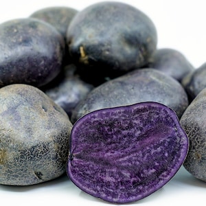 May include: A pile of dark purple potatoes, one cut in half to reveal a vibrant purple interior. The potatoes have a textured skin and are set against a white background.