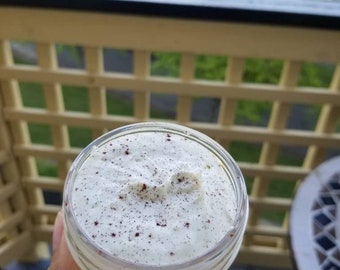 May include: A clear glass jar containing a white, creamy substance with dark brown specks. The jar is held in a hand, with a blurred outdoor background of greenery and a wooden railing.