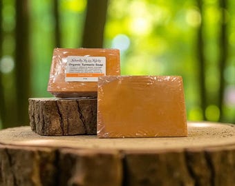 Op de afbeelding: Drie oranje biologische kurkuma zeepjes, verpakt in doorzichtig plastic, worden getoond op een houten oppervlak. Eén zeepje heeft een label met de tekst "Naturally Me By Meleky Organic Turmeric Soap Sweet Orange". De achtergrond is een vage groene bos.