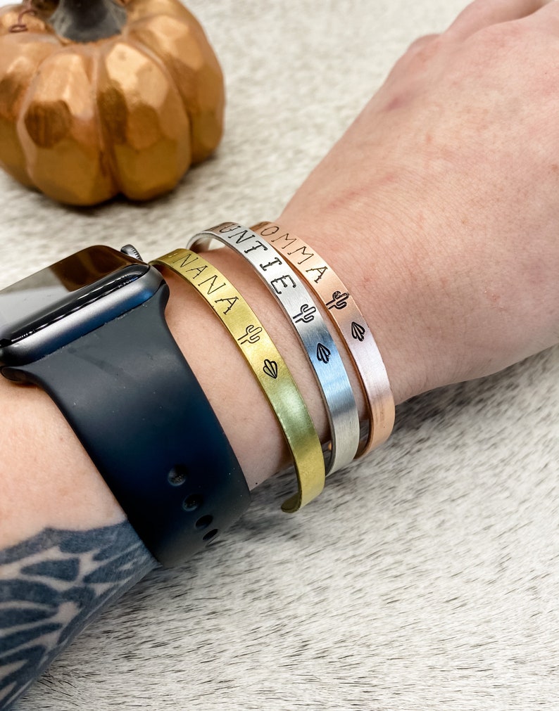 Puede incluir: Tres pulseras de oro, plata y oro rosa con los nombres grabados "NANA", "UNTIE" y "MOMMA" con un peque&ntilde;o cactus y un coraz&oacute;n.