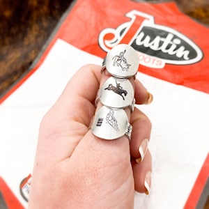 Peut inclure: Trois bagues en argent avec différents motifs de cowboy chevauchant un taureau. Les bagues sont empilées sur un doigt. Les bagues sont gravées avec les mots "Justin Boots".