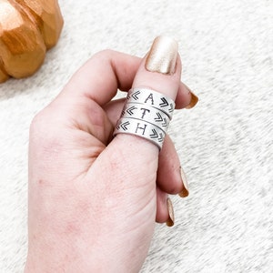 Peut inclure: Quatre anneaux en argent empilés sur un doigt, chaque anneau porte une seule lettre gravée, épeler le mot "FAITH" avec des flèches pointant dans des directions opposées sur chaque anneau.