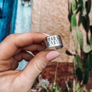 May include: A silver ring with the words "Mama Tried" engraved on it. The ring is being held by a hand with pink nail polish.