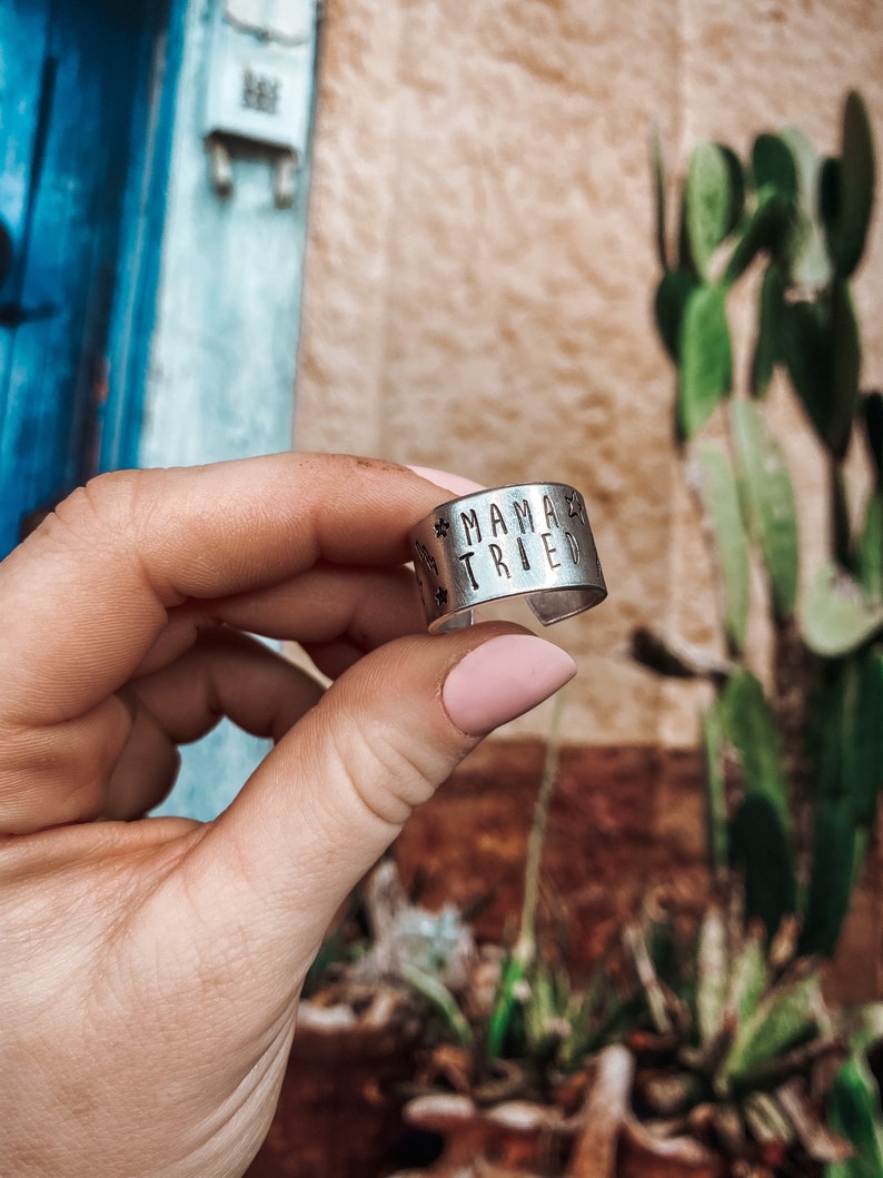 May include: A silver ring with the words "Mama Tried" engraved on it. The ring is being held by a hand with pink nail polish.