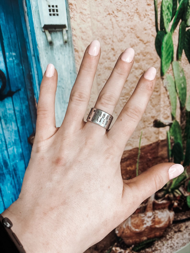 May include: A silver ring with the words "Mama'z Tried" engraved on it. The ring is worn on the middle finger of a hand with light pink nail polish.
