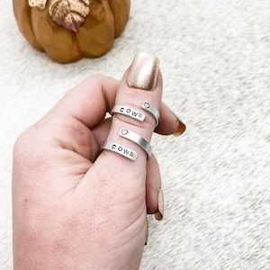 May include: Two silver rings with the word "COWS" and a small heart on each ring. The rings are worn on a person's thumb.