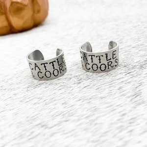 May include: Two silver metal rings with the words "Cattle & Coors" engraved on them.