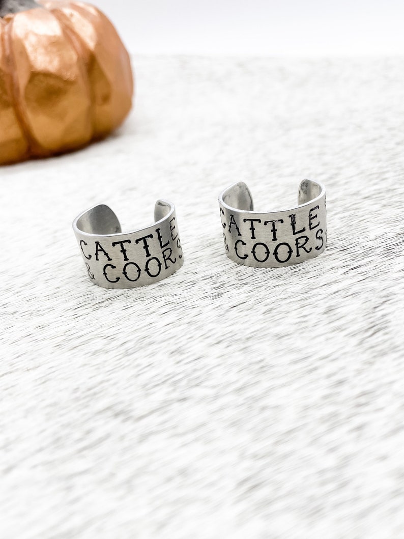 May include: Two silver metal rings with the words "Cattle & Coors" engraved on them.