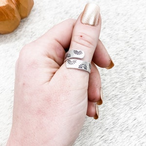 May include: A silver adjustable ring with a sunburst design. The ring is worn on a person's thumb.