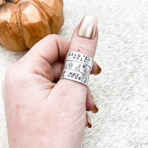 May include: A set of four silver thumb rings with different engraved sayings. The rings are stacked on a person's thumb. The top ring says "Give 'em Hell". The second ring has a cactus design. The third ring says "Hot Mess". The bottom ring has a cactus design.