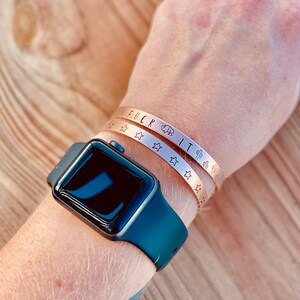 May include: Two gold-toned bracelets on a person's wrist. The top bracelet has the words "F*CK IT" engraved on it. The bottom bracelet has a repeating pattern of stars and cacti.