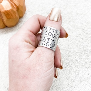May include: Two silver rings stacked on a finger. The rings are engraved with the text "Cattle & Coors".