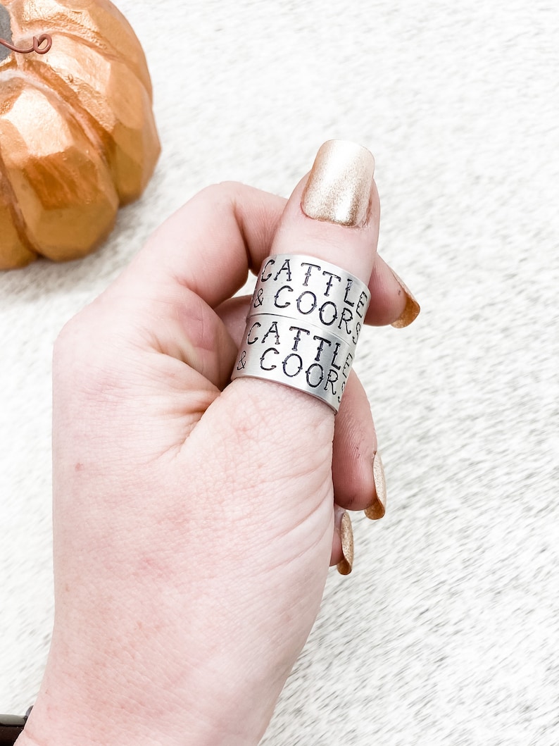 May include: Two silver rings stacked on a finger. The rings are engraved with the text "Cattle & Coors".