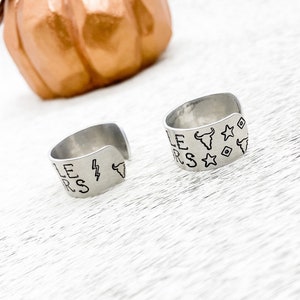May include: Two silver rings with engraved designs. One ring has the words "Little" and "Stars" engraved on it, along with a lightning bolt and a bull's head. The other ring has the words "Little" and "Stars" engraved on it, along with a bull's head and stars.