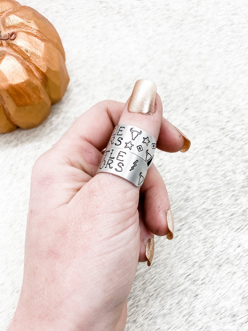 May include: Two silver rings with the words "Little Stars" and "Little Riders" engraved on them. The rings are worn on a person's thumb.