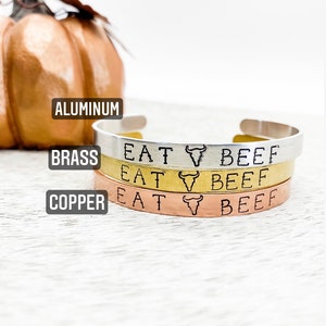May include: Three stacked bracelets in aluminum, brass, and copper. Each bracelet has the text "EAT BEEF" with a bull's head symbol.